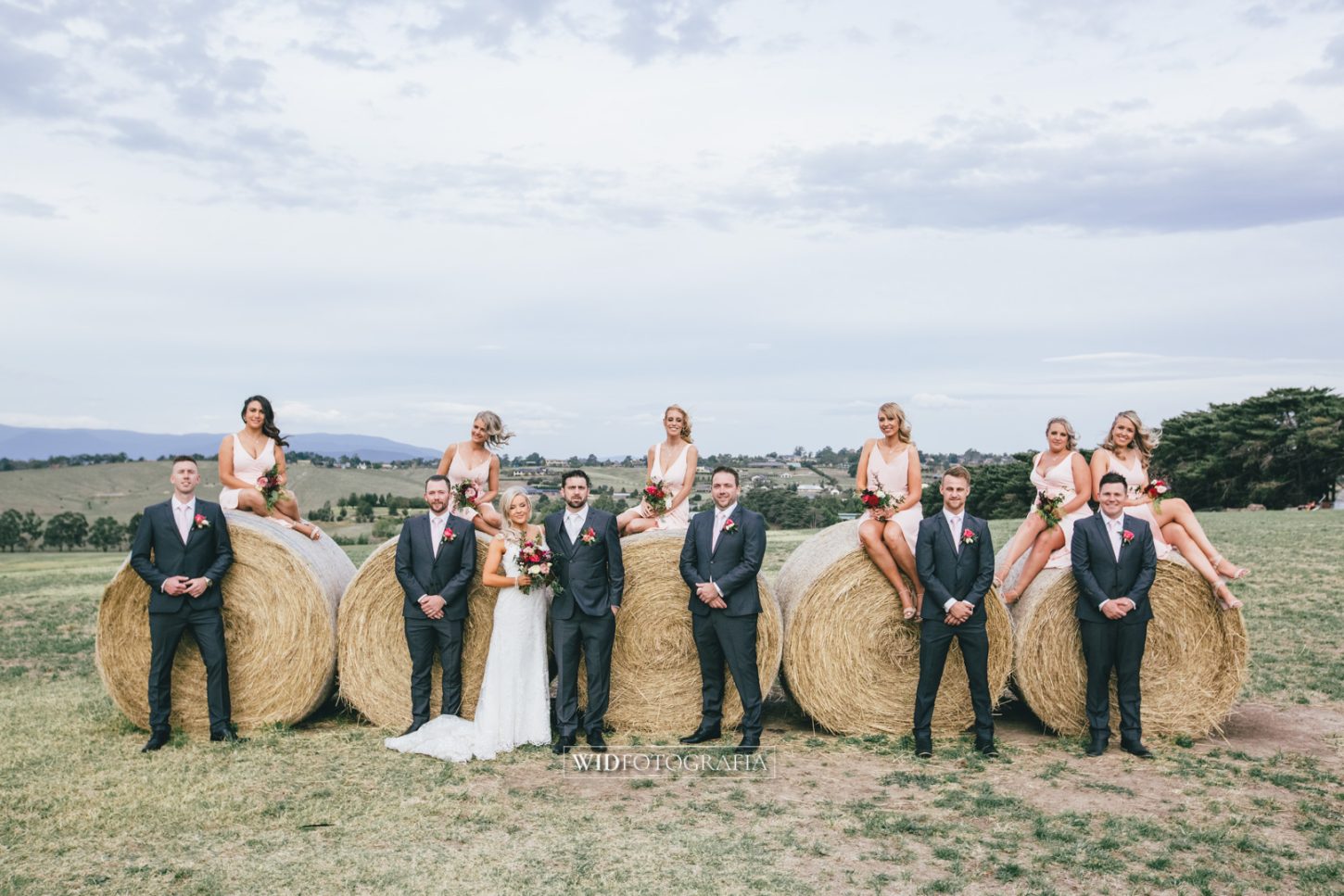 Wedding photo of Bec & Ryan and their bridesmaids and groomsmen, posing on stacks of hay at Vue on Halcyon