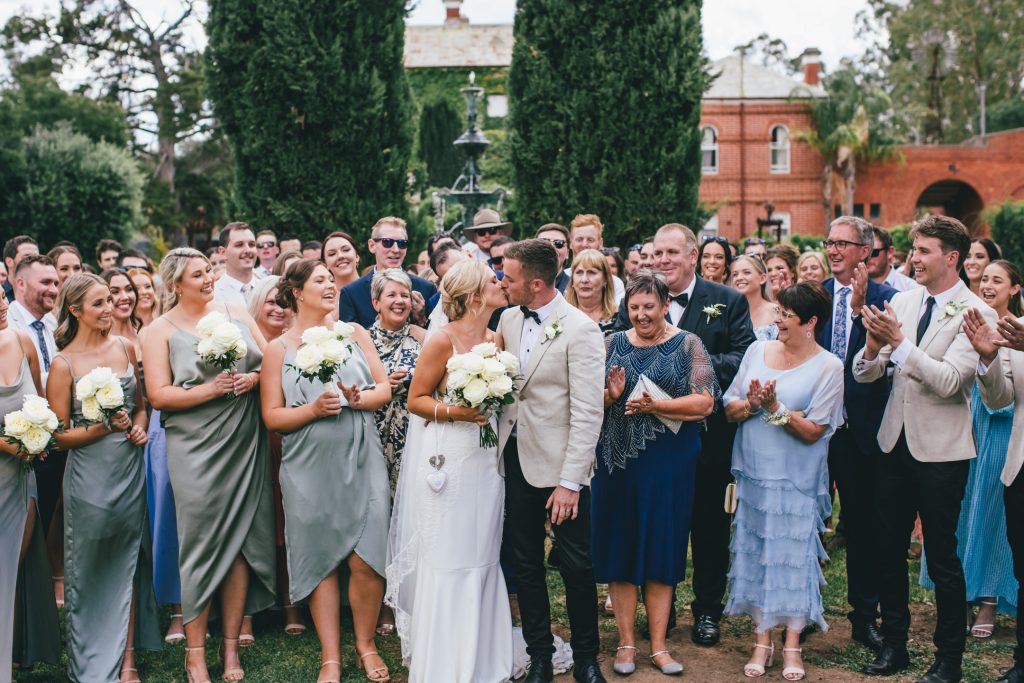 Group photo of bride and groom kissing and guests cheering at Ravenswood Homestead Wedding.