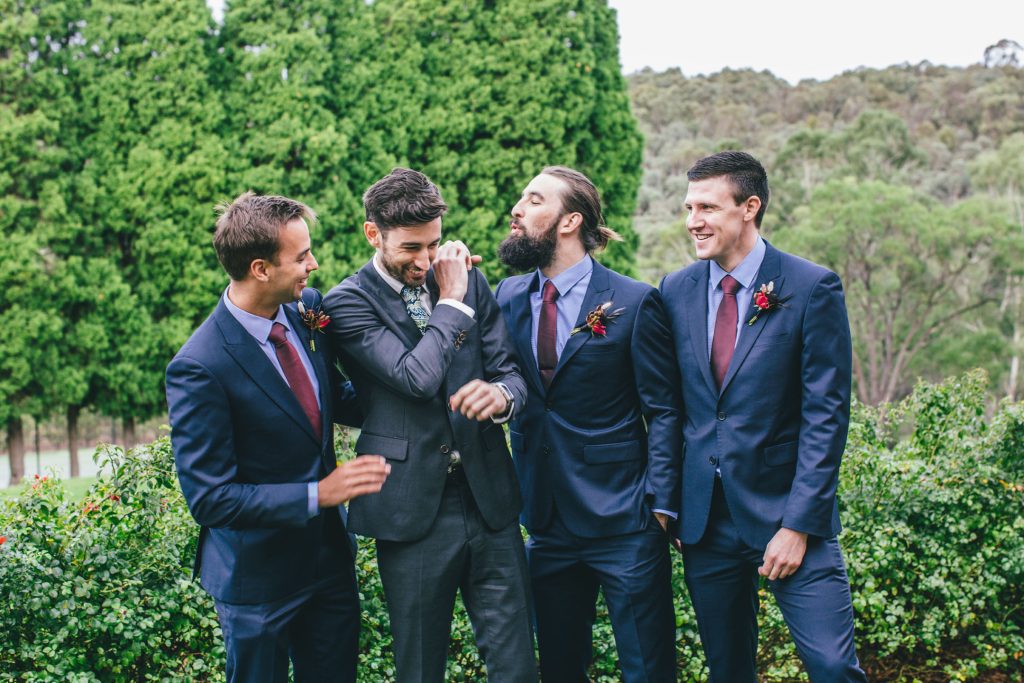 Groom and his groomsmen joking in a garden wearing suits for Farm Vigano South Morang wedding.