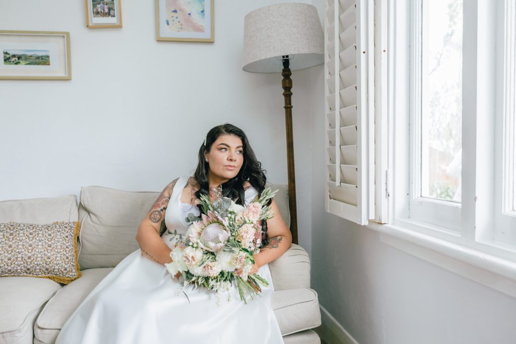 Bride sitting on the couch holding bouquet of flower while looking at outside the window.