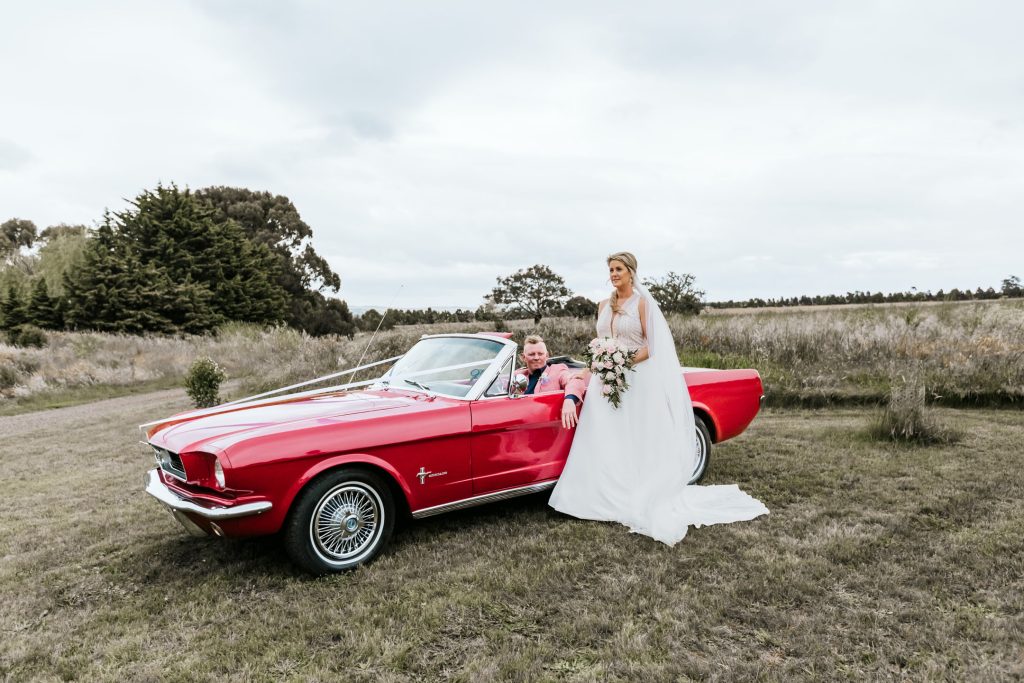 Bride standing while groom sitting inside the red mustang wedding car.